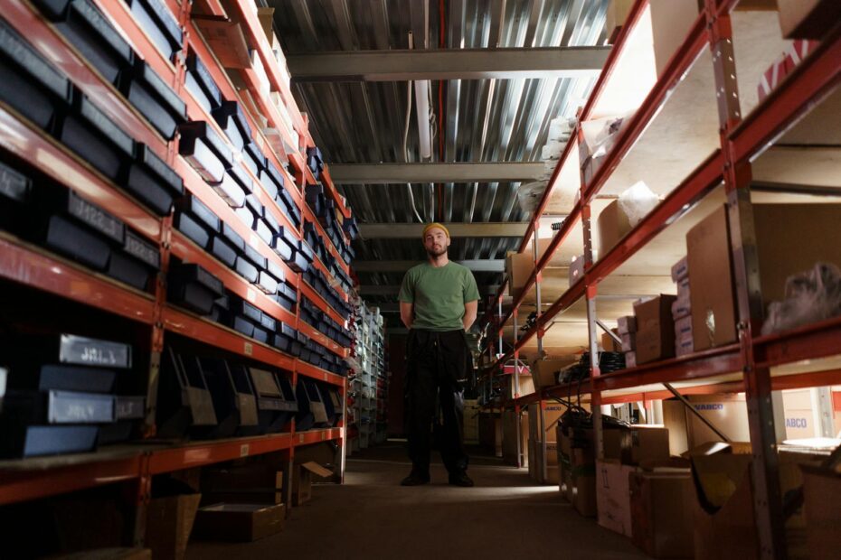 A man standing in a warehouse aisle surrounded by shelves filled with boxes and inventory.